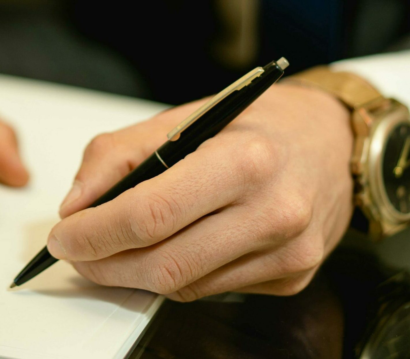 Hand writing with pen on paper. A gold watch is visible.
