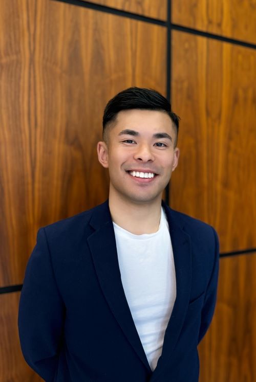 Smiling man in a navy blazer and white t-shirt stands against a wood-paneled wall.