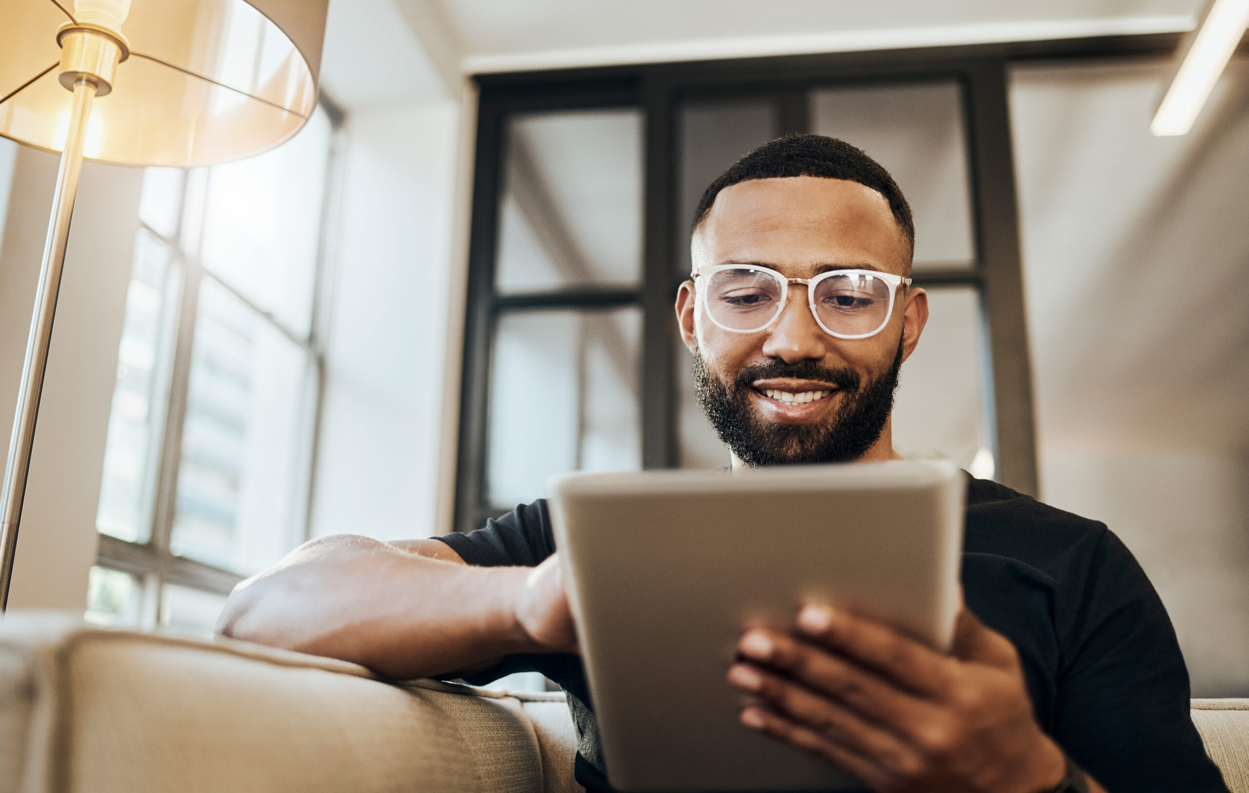 Smiling man in glasses sitting on a sofa, using a tablet in a bright modern living room.